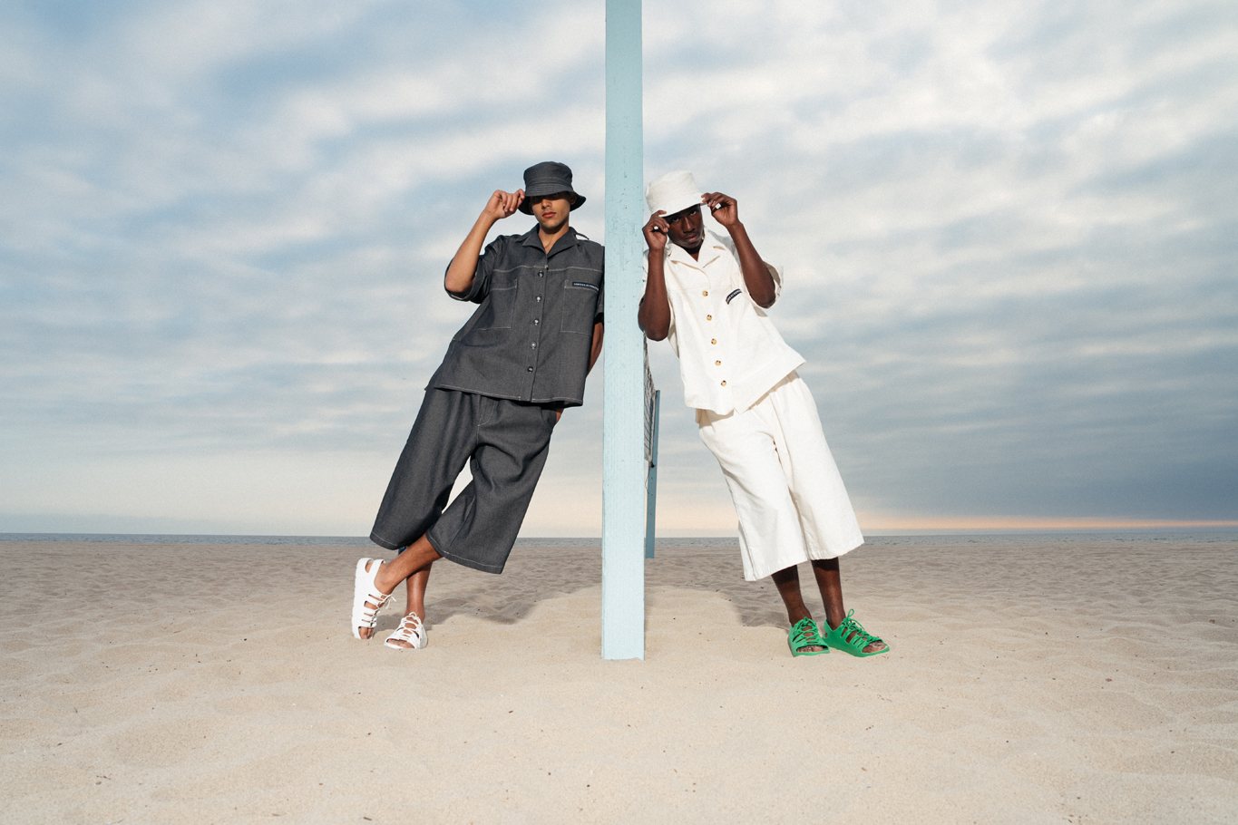 Two models in matching bucket-hat denim and off-white sets beside a beach volleyball pole