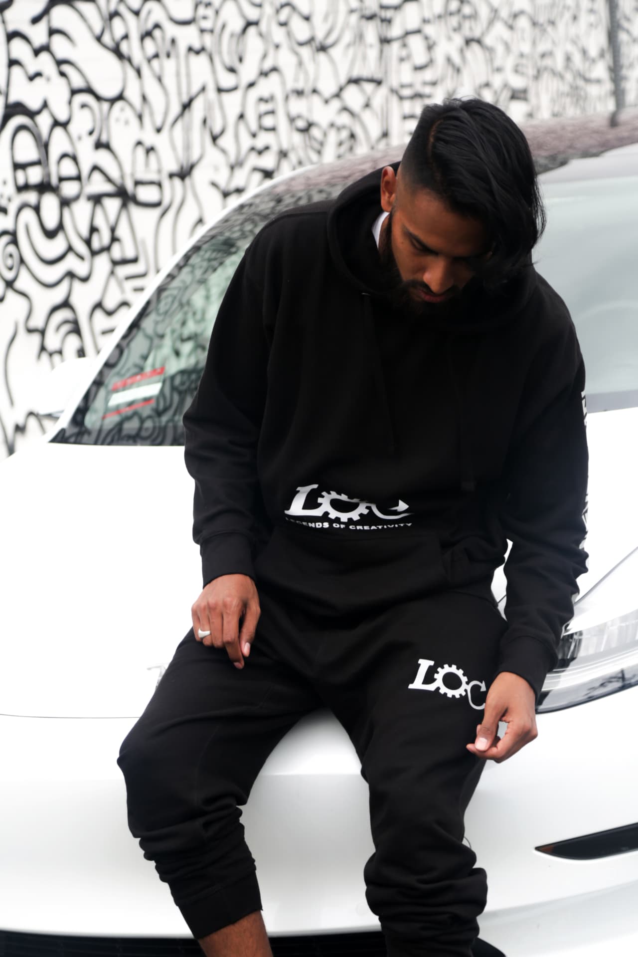 Black hooded set photographed against a graphic mural and white car